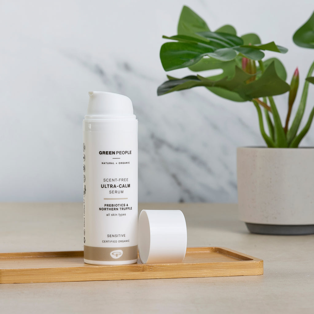 A white bottle of Scent Free Ultra Calm Serum rests on a small bamboo tray next to a tropical green palm leaf.