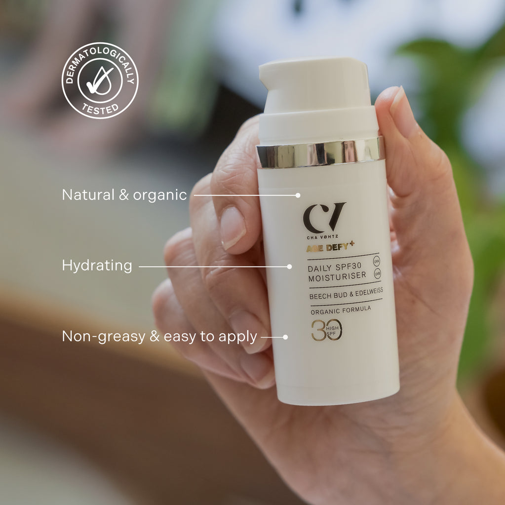 Hand holding a white pump bottle of moisturizer with silver trim, angled left with blurred green and brown background.