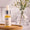 A glass dropper bottle with a yellow liquid, branded label, on a wooden tray, with a flowered vase behind.